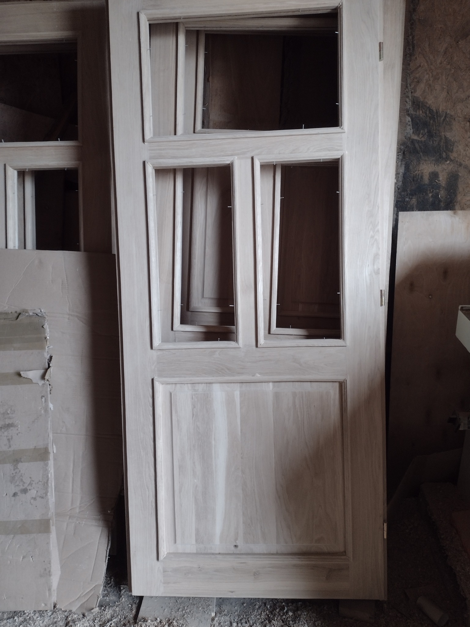 Surowa, drewniana rama okienna z trzema przeszkleniami, częściowo oszklona, oparta o ścianę w warsztacie, z widocznymi śladami obróbki drewna.