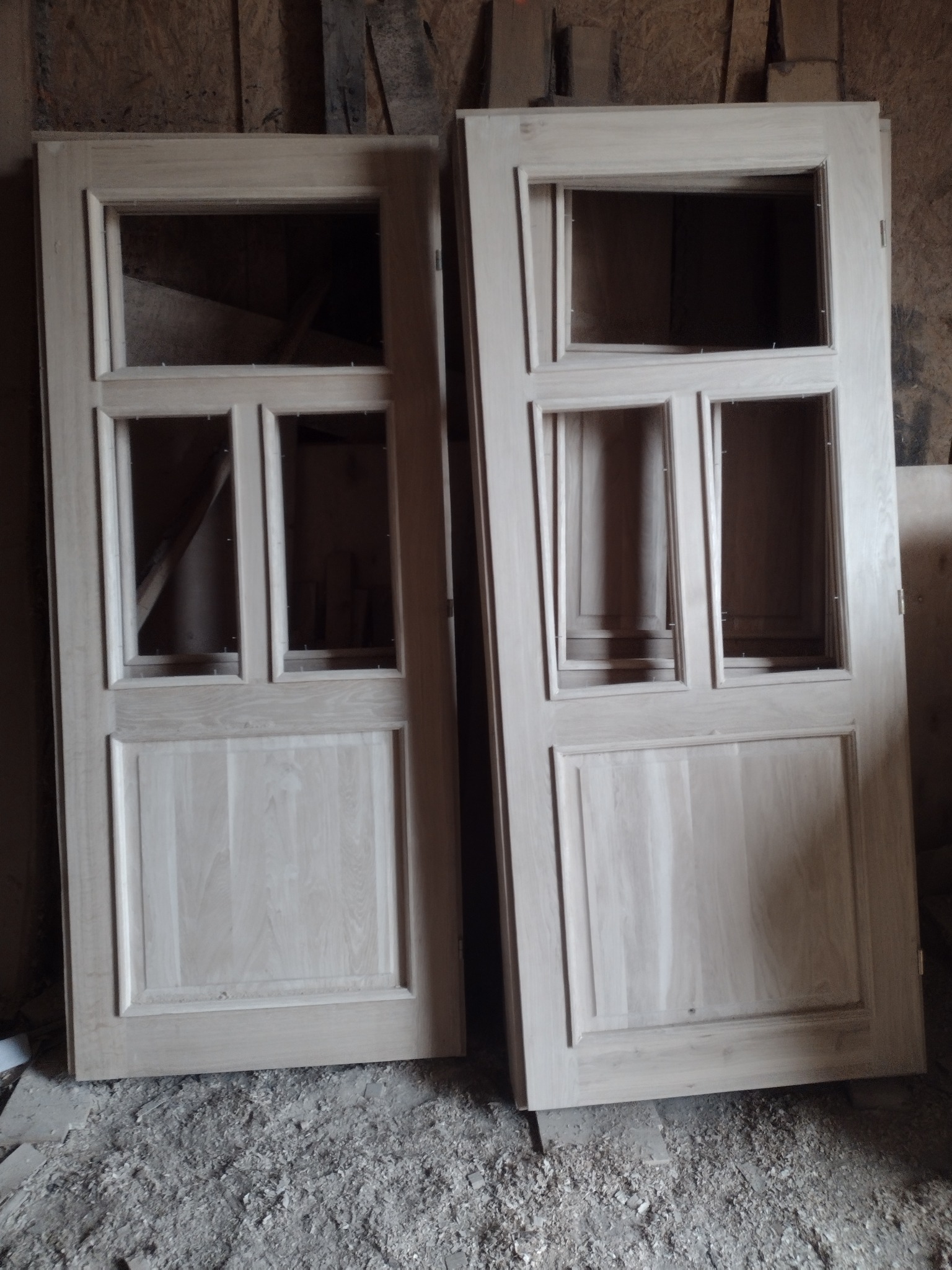 Dwie surowe, drewniane ramy okienne z podziałami, o jasnym odcieniu, stojące na podłodze pokrytej wiórami i pyłem drzewnym, w warsztacie.