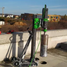 Wiertnica Eibenstock na dachu budynku w trakcie budowy, w tle widoczne budynki mieszkalne i dźwigi budowlane, obok leżą słuchawki ochronne i puszka WD-40.