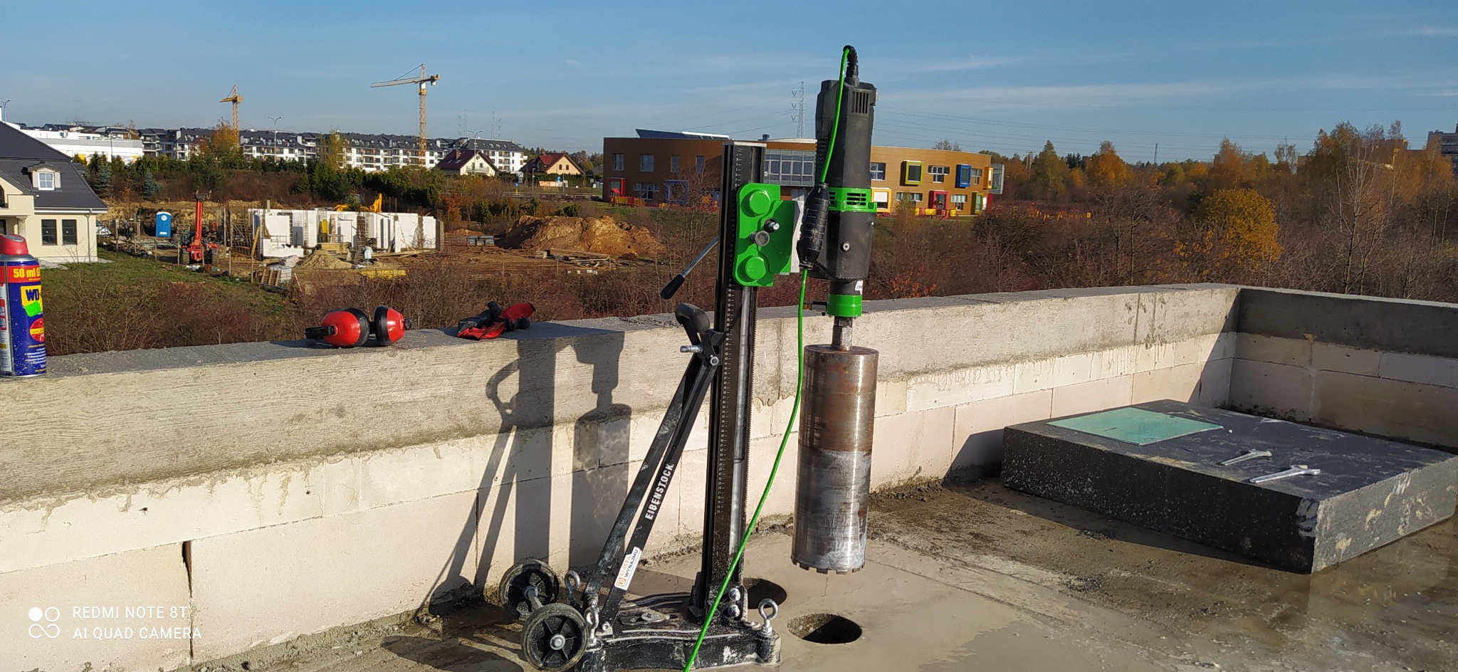 Wiertnica Eibenstock na dachu budynku w trakcie budowy, w tle widoczne budynki mieszkalne i dźwigi budowlane, obok leżą słuchawki ochronne i puszka WD-40.