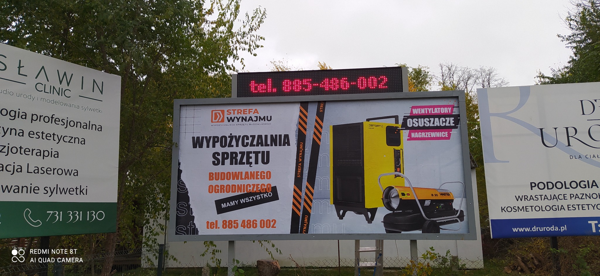Billboard reklamujący wypożyczalnię sprzętu budowlanego, z widocznymi wentylatorami, osuszaczami i nagrzewnicami oraz numerem telefonu, obok reklamy kliniki urody i gabinetu podologicznego.