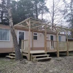 Mobilny domek z dobudowanym drewnianym tarasem i pergolą, widok na surową konstrukcję w otoczeniu drzew.