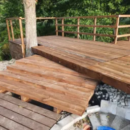 Drewniany taras z balustradą, schodami i elementami kamiennymi, otoczony zielenią, z drzewem przechodzącym przez konstrukcję.