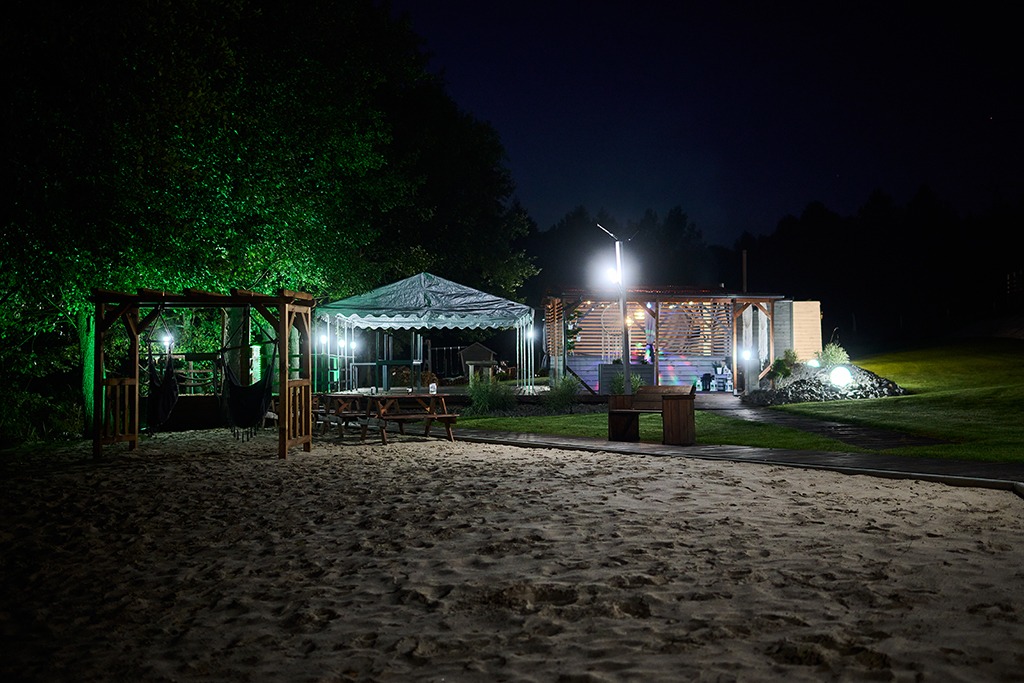 Oświetlony nocą teren rekreacyjny z piaskiem, altaną z grillem, zadaszeniem i drewnianą konstrukcją z wiszącymi hamakami.