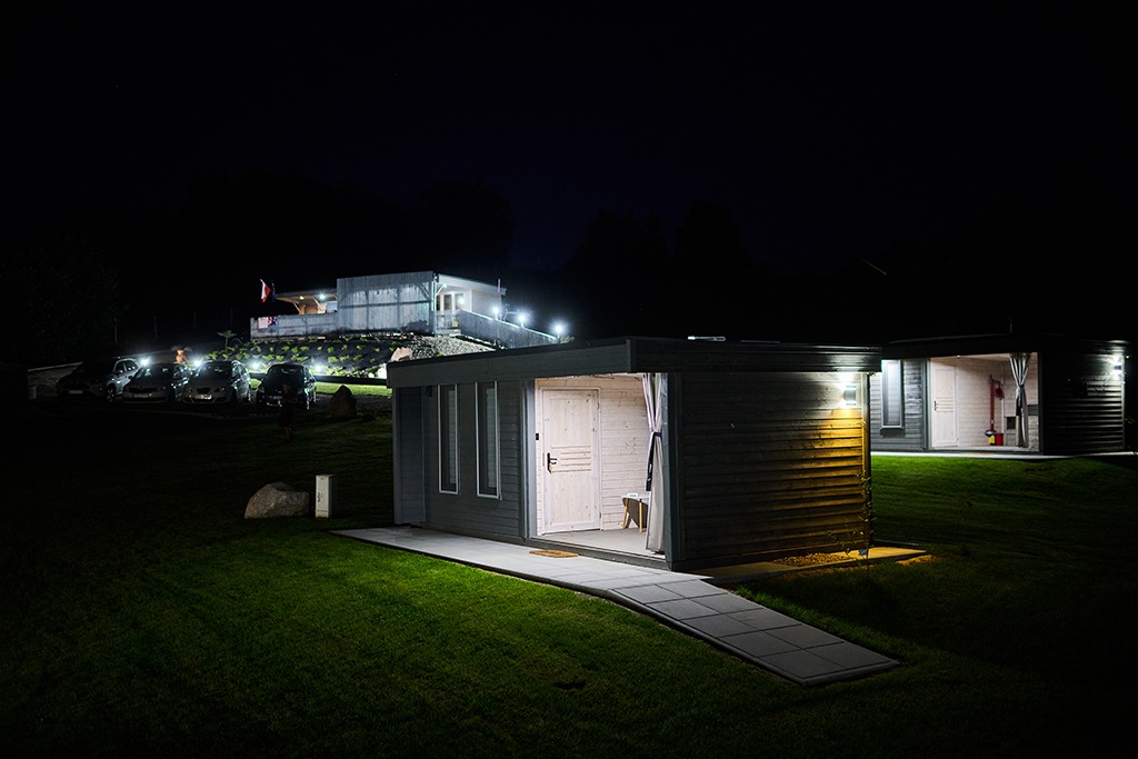 Dwa nowoczesne, drewniane domki letniskowe oświetlone nocą, z widoczną zieloną trawą i budynkiem w tle, oświetlonym reflektorami i z powiewającą flagą.