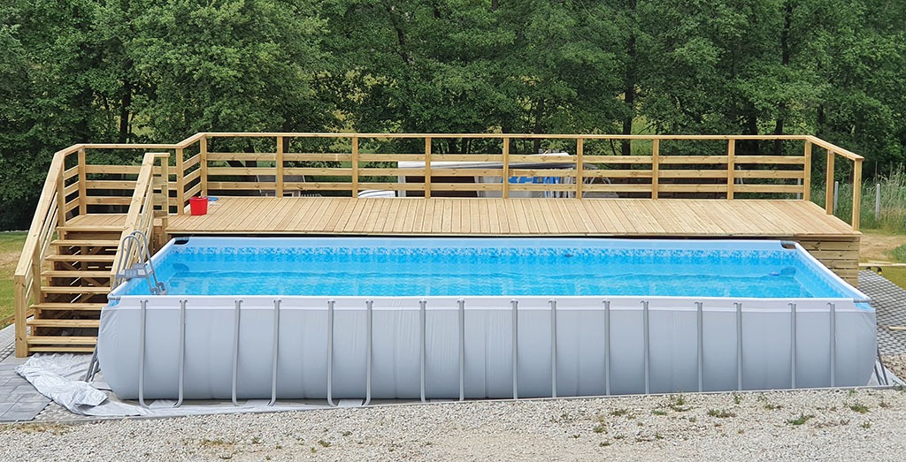Prostokątny basen naziemny z metalową ramą i drewnianym podestem z barierkami i schodami, otoczony zielenią.