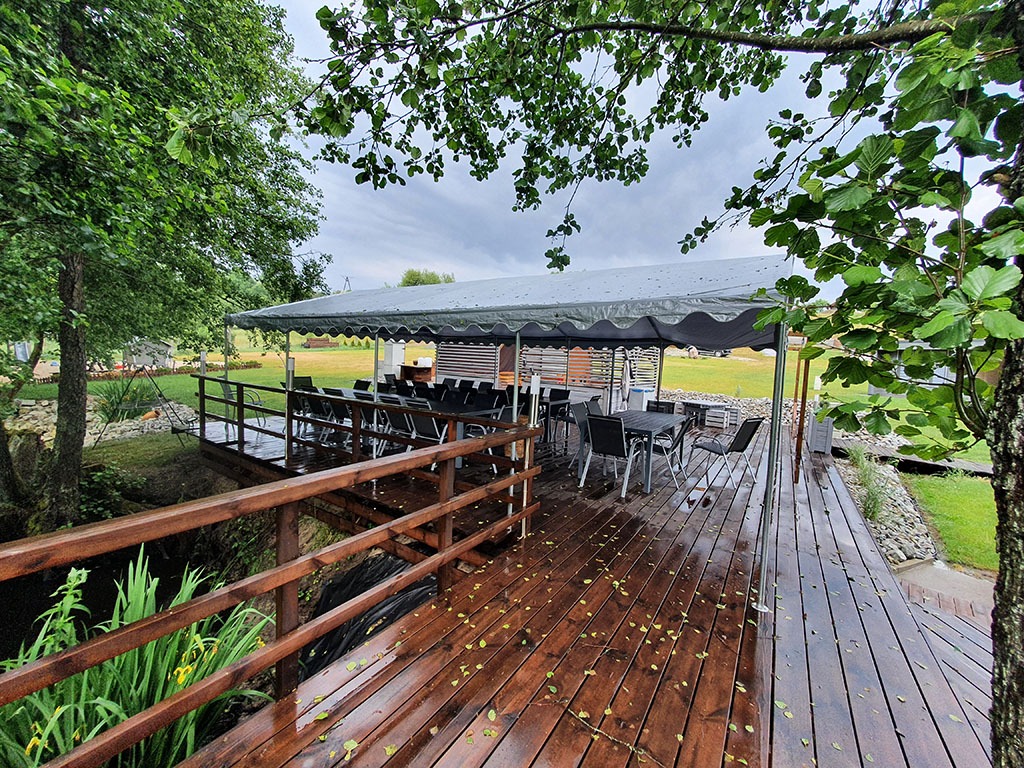 Zadaszony taras z drewnianą podłogą nad strumieniem, z widokiem na zielony trawnik i drzewa w tle, częściowo pokryty opadłymi liśćmi.