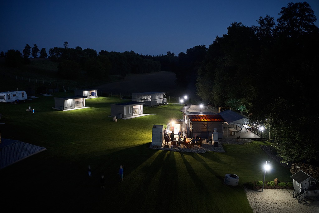 Nocny widok na ośrodek wypoczynkowy z domkami modułowymi, oświetlonym terenem, miejscem na ognisko i placem zabaw.
