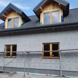 Ocieplony dom z szarym tynkiem w kropki, drewnianymi oknami i czarnym dachem z dachówką, w trakcie prac elewacyjnych, widoczne rusztowania.