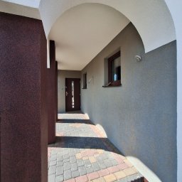 MAX-ECO - Zadaszone wejście do budynku z brązowymi drzwiami i oknem. Elewacja w odcieniach szarości i brązu, wykończona kostką brukową. Widoczne oświetlenie zewnętrzne.