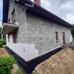 MAX-ECO - Ocieplanie fragmentu ściany domu z szarego bloczka, widoczna czarna folia fundamentowa i biały styropian. Częściowo ocieplona elewacja w trakcie prac.