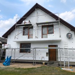 MAX-ECO - Dom jednorodzinny w trakcie ocieplania styropianem, widoczne rusztowania i zabezpieczone okna folią. Częściowo ocieplona elewacja, widoczne puszki na elewacji.