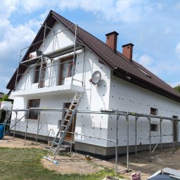 MAX-ECO - Dom w trakcie ocieplania styropianem, widoczne rusztowania i drabina. Brązowy dach, białe ściany, błękitne niebo. Etap prac z widocznymi elementami montażowymi.