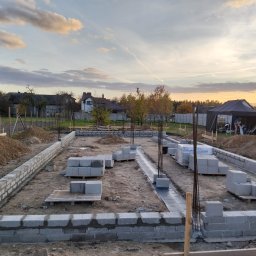 MAX-ECO - Fundamenty domu jednorodzinnego w trakcie budowy, z widocznymi bloczkami betonowymi i zbrojeniami, na tle zachodzącego słońca i zabudowy jednorodzinnej.