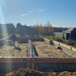 MAX-ECO - Fundamenty domu jednorodzinnego w trakcie budowy. Widoczne zbrojenia, szalunki i pracujący robotnicy. W tle zabudowa jednorodzinna i błękitne niebo.