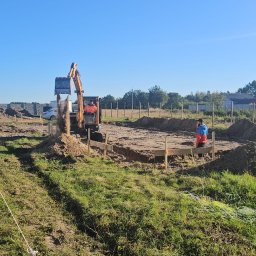 MAX-ECO - Prace ziemne pod budowę domu jednorodzinnego: koparka wyrównuje teren pod fundamenty. Widoczne drewniane szalunki i pracownik w oddali. Błękitne niebo.