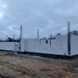 MAX-ECO - Budowa domu z białych bloczków H+H, widoczne zbrojenia pionowe. Prace murarskie w toku, teren budowy z błotnistym podłożem, pochmurne niebo w tle.