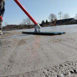 MAX-ECO - Wyrównywanie wylewki betonowej na budowie domu jednorodzinnego. Widoczny pracownik z narzędziem, świeży beton i zbrojenia w tle. Słoneczny dzień.
