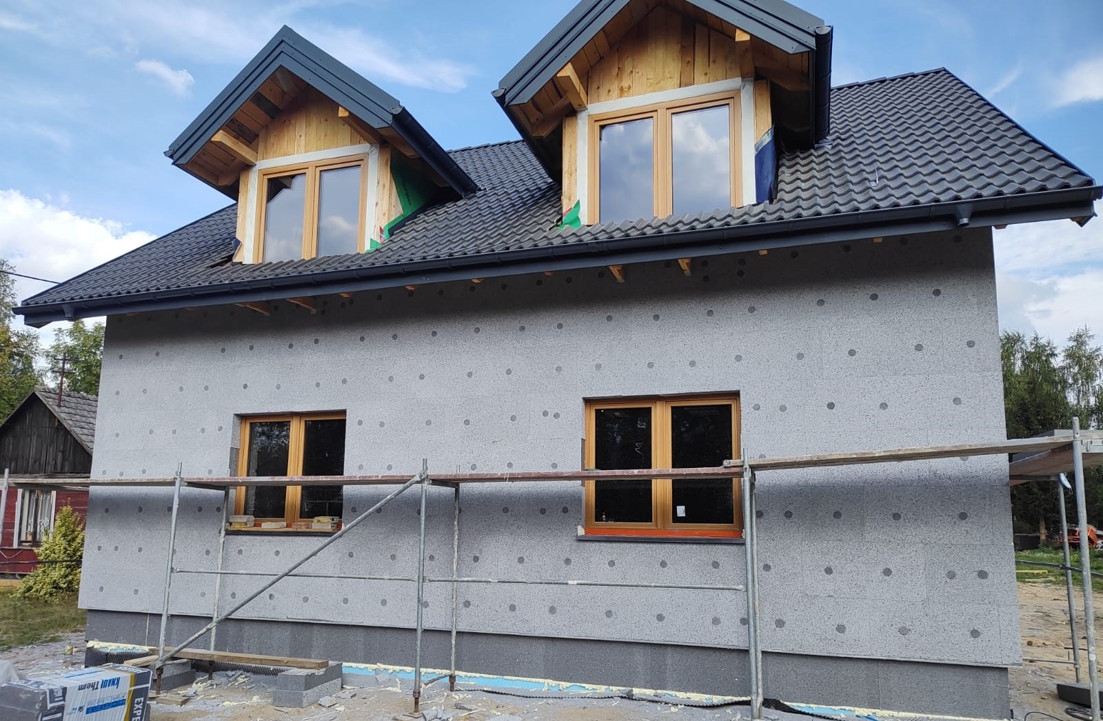 Ocieplony dom z szarym tynkiem w kropki, drewnianymi oknami i czarnym dachem z dachówką, w trakcie prac elewacyjnych, widoczne rusztowania.