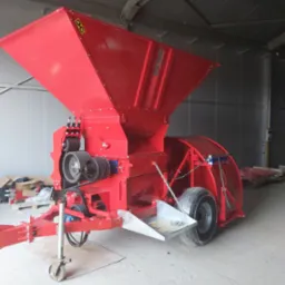 Czerwona, mobilna maszyna do rozdrabniania gałęzi z dużym zasobnikiem, widoczna w hali produkcyjnej, z elementami zabezpieczonymi folią.