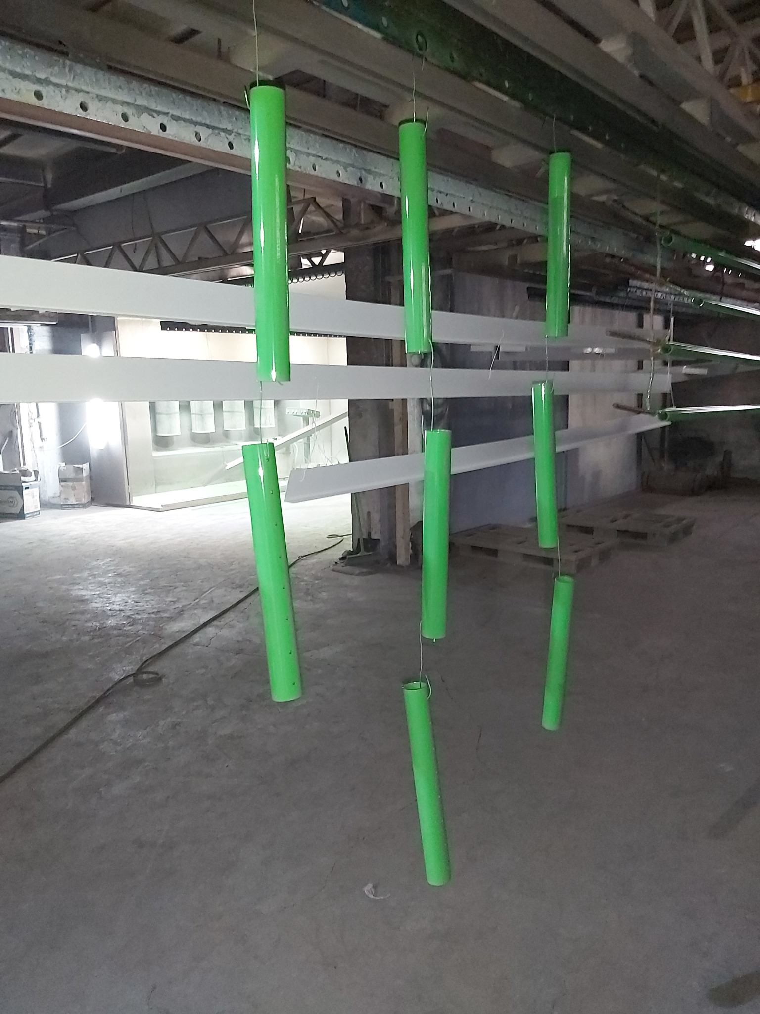 Metalowe rury w jaskrawozielonym kolorze zawieszone na metalowych hakach, prawdopodobnie po procesie malowania, w przemysłowym otoczeniu.