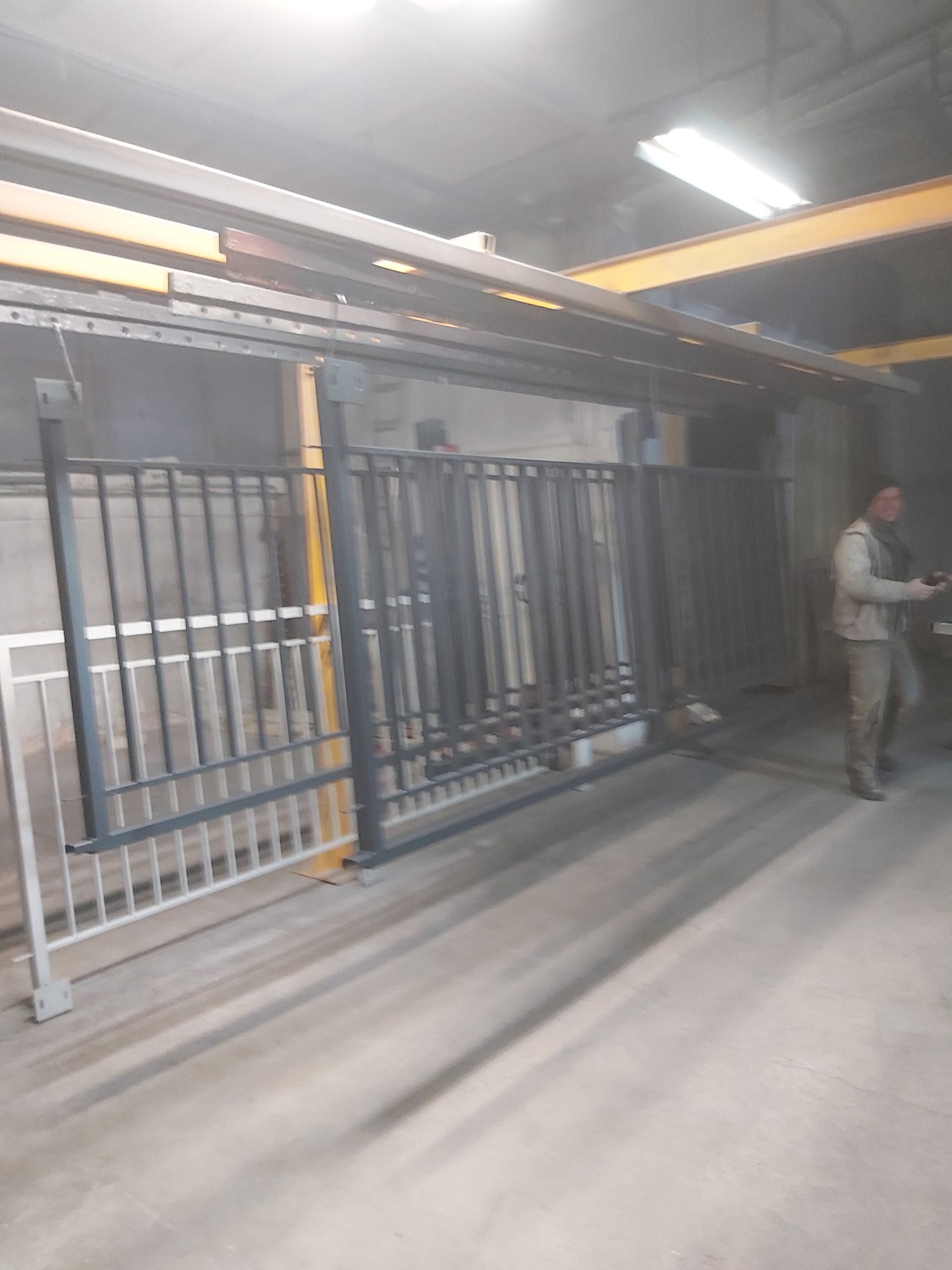 Metalowe elementy ogrodzenia, częściowo pomalowane na biało, zawieszone na konstrukcji przed procesem malowania, z widoczną osobą w tle.