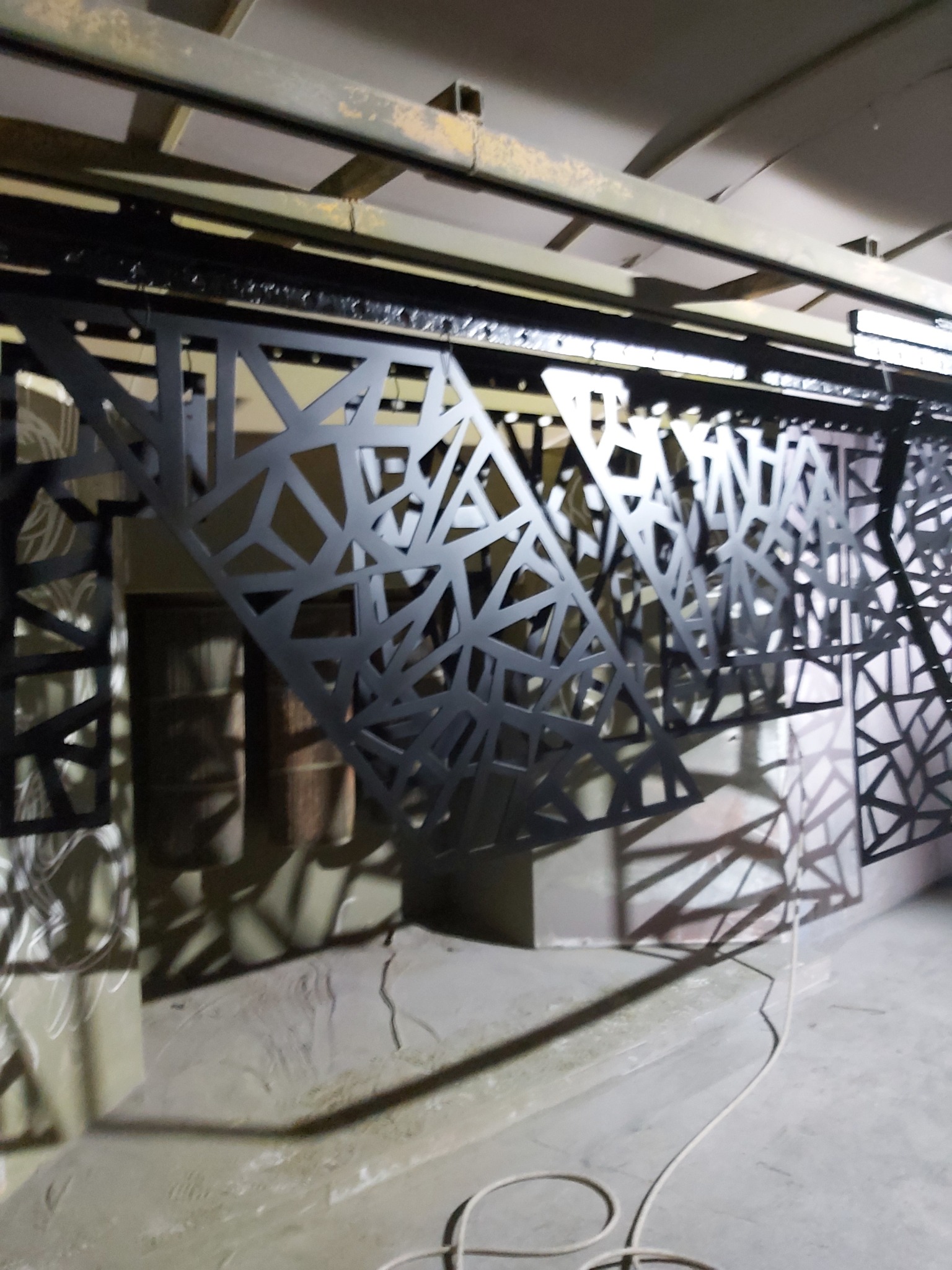 Metalowe panele dekoracyjne z geometrycznym wzorem, zawieszone na metalowej konstrukcji, prawdopodobnie w trakcie procesu wykończeniowego, rzucające cienie na betonową podłogę.