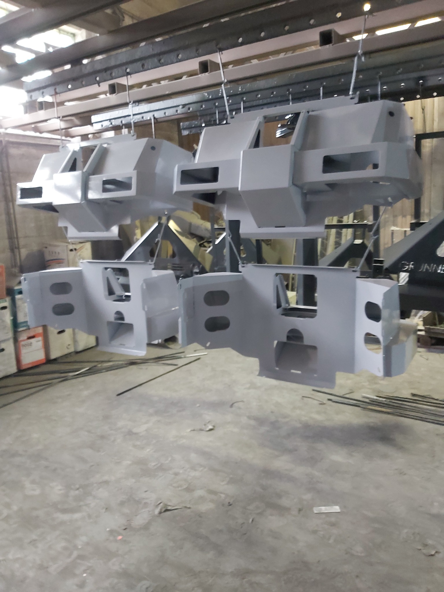 Metalowe elementy konstrukcyjne zawieszone na hakach, pomalowane na szaro, w hali produkcyjnej. Widoczne ślady użytkowania na podłodze i elementy konstrukcyjne sufitu.