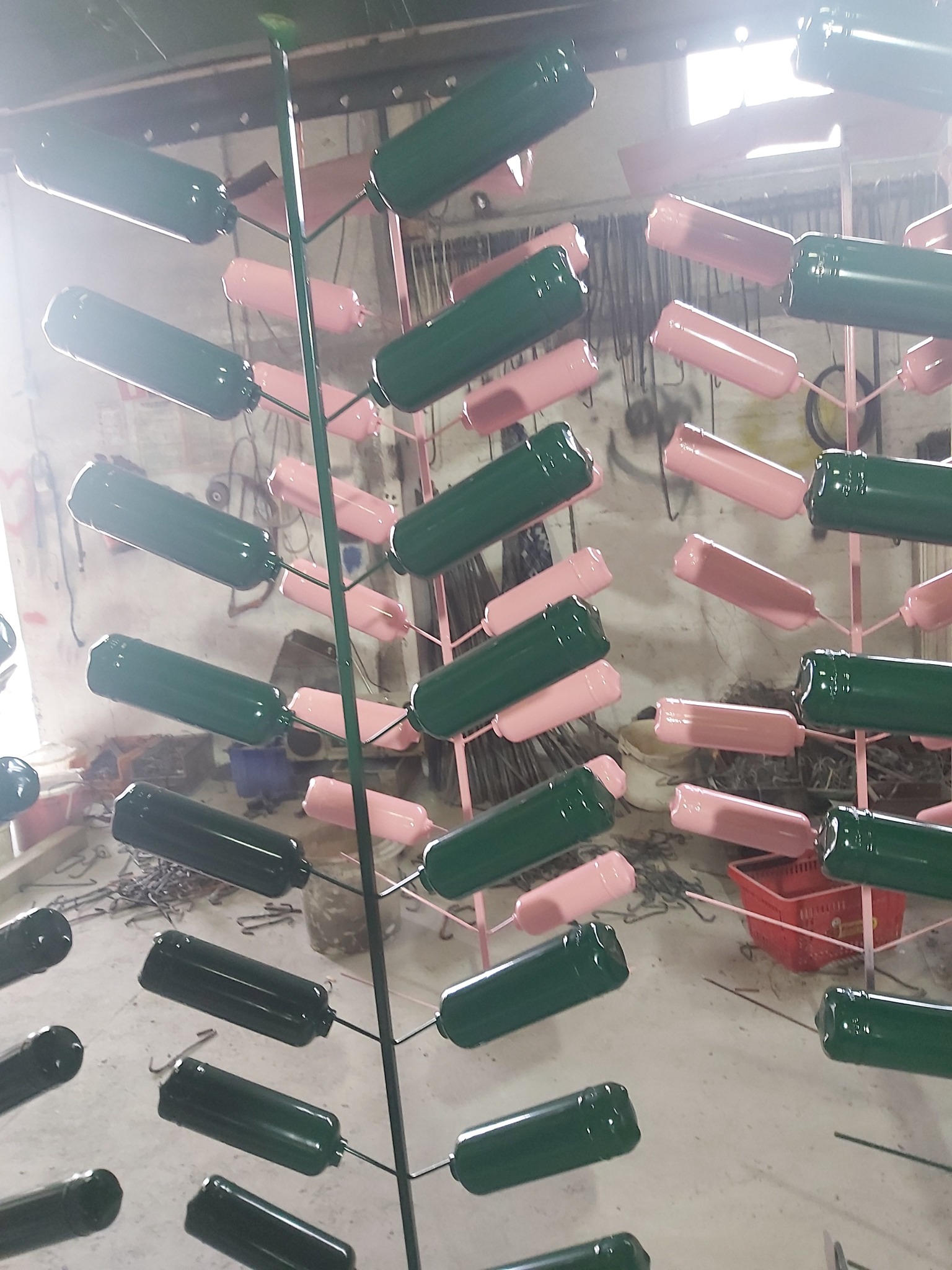 Metalowe butle zawieszone na stojakach, niektóre pomalowane na zielono, inne na różowo, w warsztacie z widocznymi narzędziami i elementami wyposażenia.