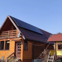 Drewniany dom z panelami fotowoltaicznymi na dachu, obok kolorowy plac zabaw dla dzieci w słoneczny dzień.