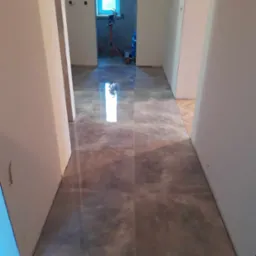 Korytarz z nową, szarą, błyszczącą podłogą z płytek, widoczne przygotowania do dalszych prac wykończeniowych w tle.