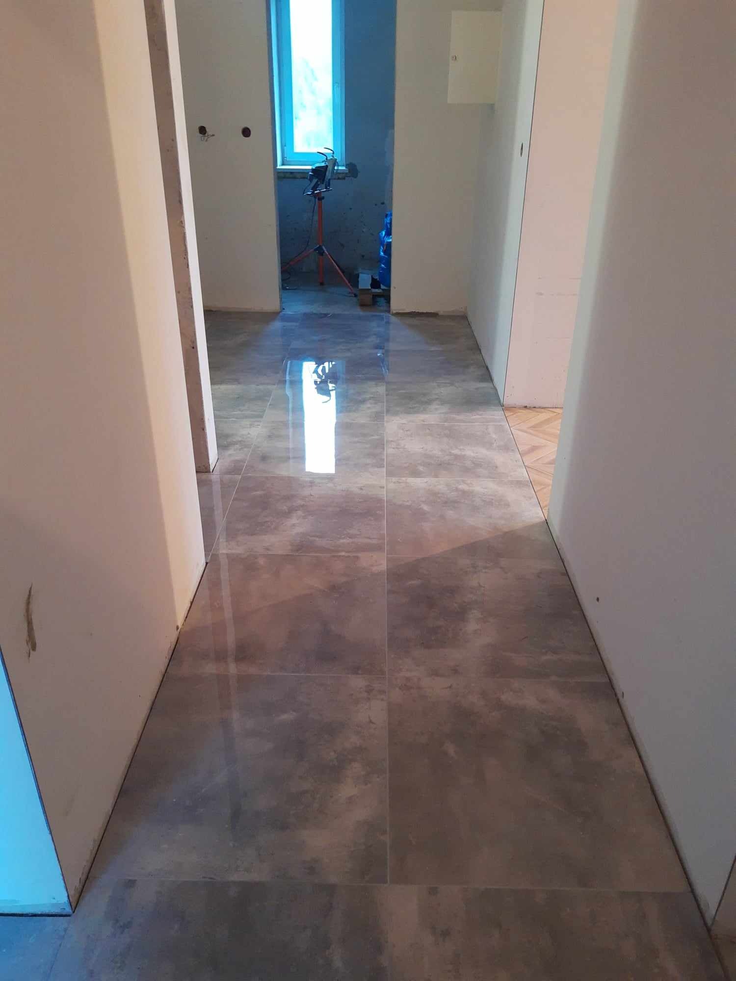 Korytarz z nową, szarą, błyszczącą podłogą z płytek, widoczne przygotowania do dalszych prac wykończeniowych w tle.