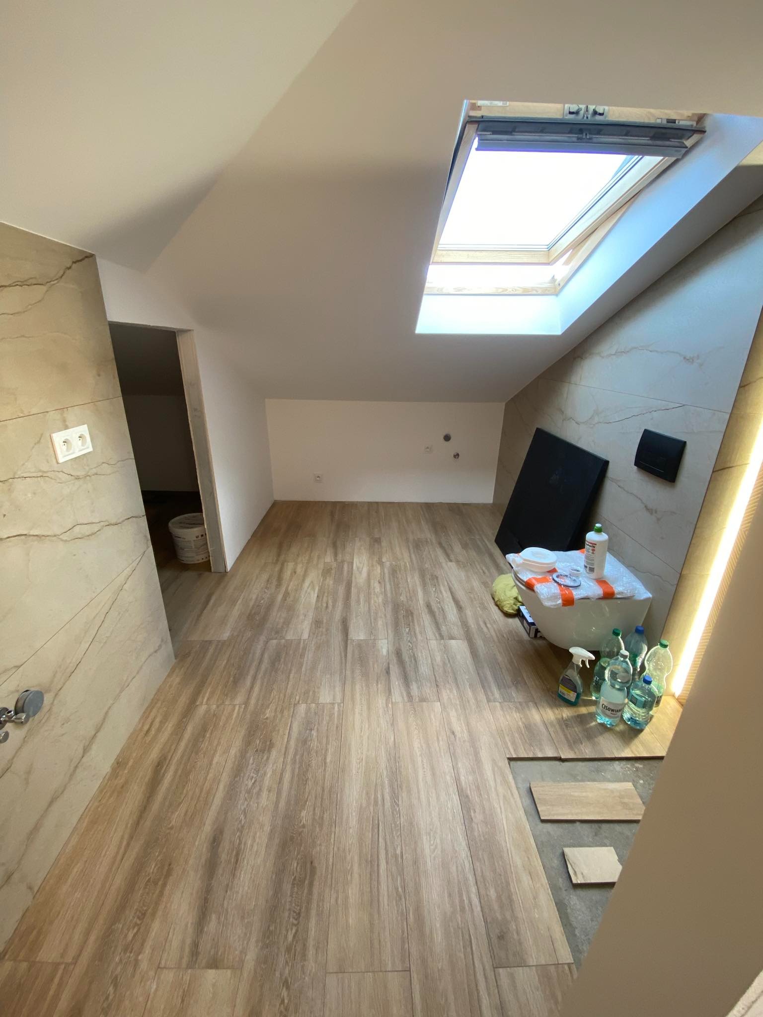 Ukończona łazienka na poddaszu z drewnopodobnymi płytkami podłogowymi, częściowo zakryta folią i materiałami budowlanymi, z widocznym oknem dachowym i częściowo wykończoną ścianą z płytek...