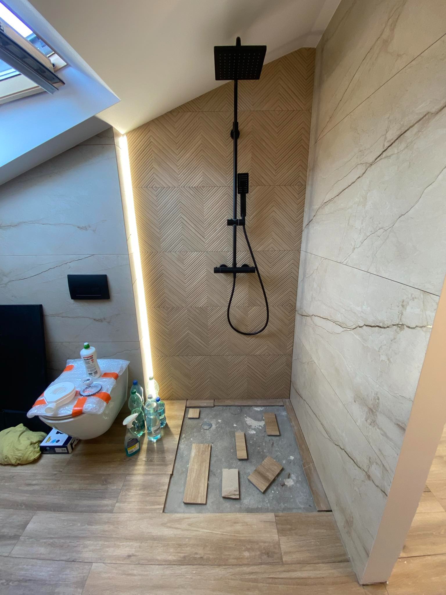 Ukośna łazienka w trakcie wykańczania: montaż płytek podłogowych w strefie prysznica z czarną deszczownicą, widoczne materiały i chemia budowlana, podświetlenie LED wzdłuż ściany.