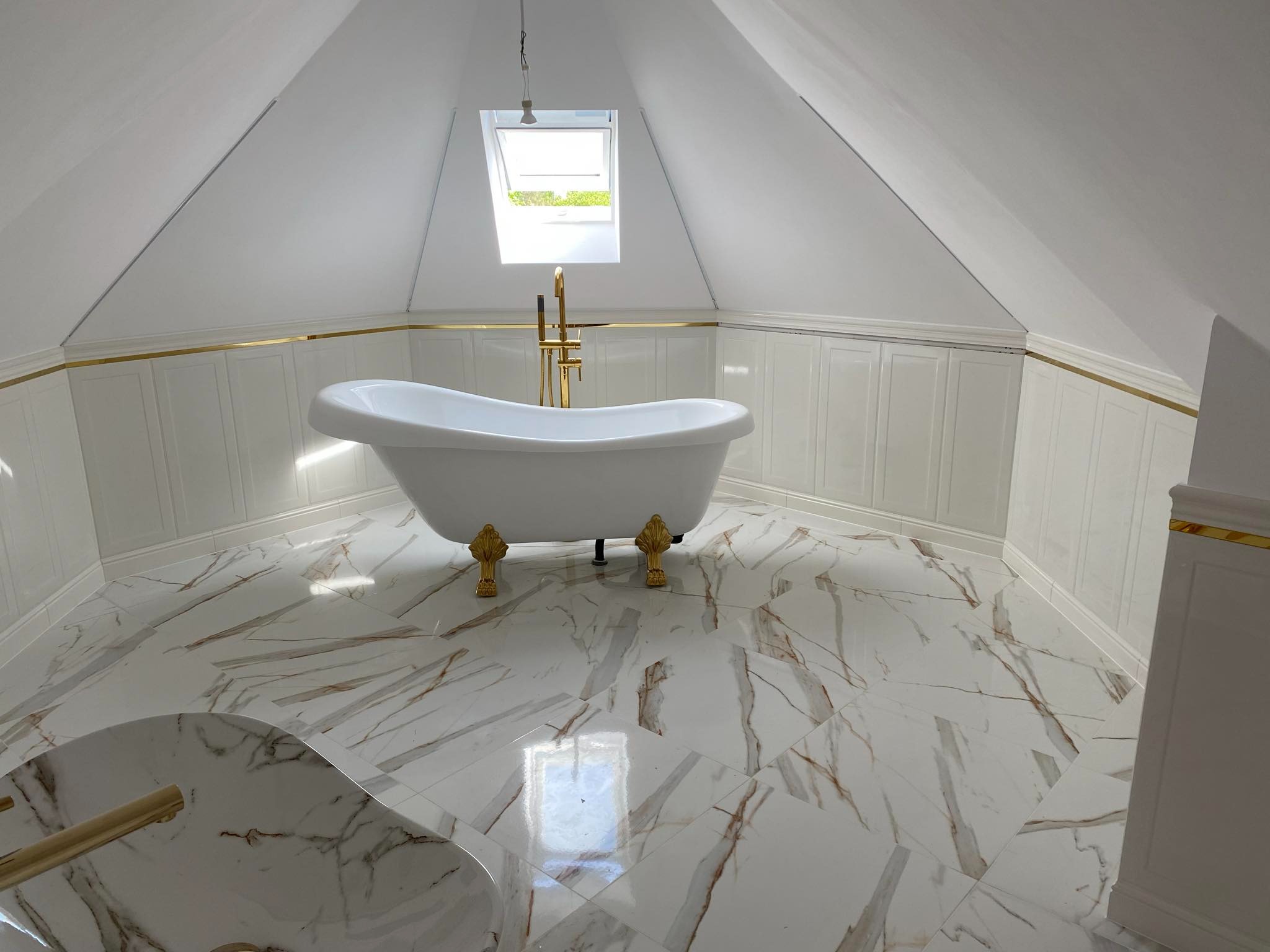 Biała wanna wolnostojąca na złotych nóżkach w łazience na poddaszu z marmurową podłogą i oknem dachowym, złote akcenty dekoracyjne na ścianach.