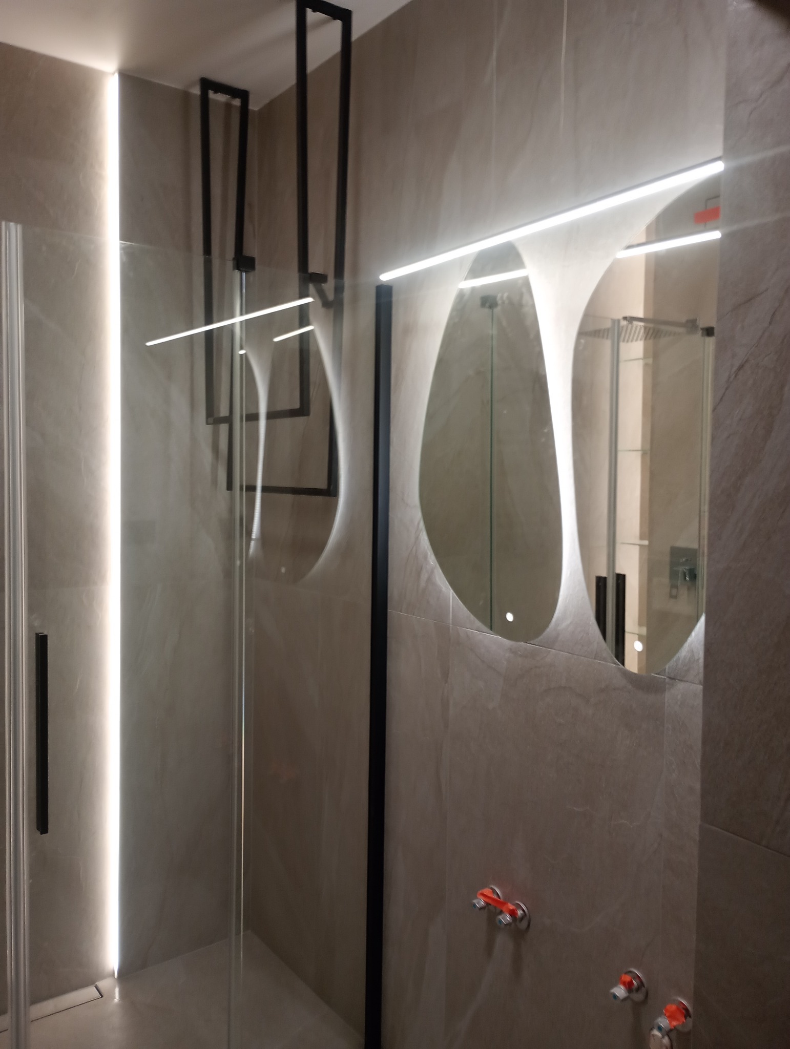 Nowoczesna łazienka z kabiną prysznicową typu walk-in, podświetlanym lustrem o organicznym kształcie i szarą, matową glazurą.