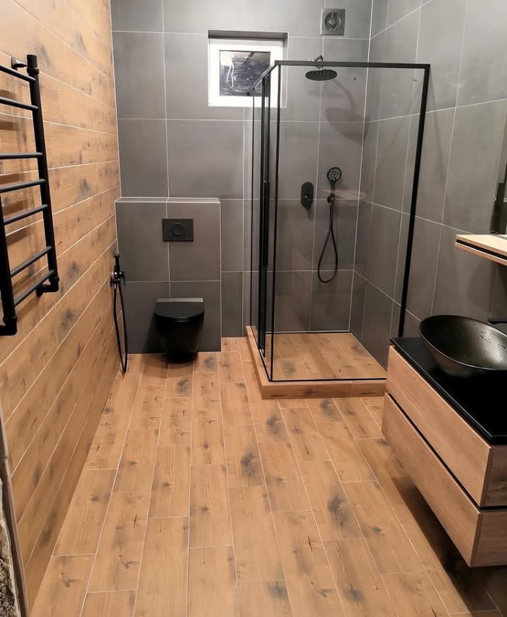 Nowoczesna łazienka z kabiną prysznicową typu walk-in z czarnymi profilami, drewnopodobnymi płytkami na podłodze i ścianie, szare płytki na ścianach, czarna armatura i toaleta wisząca,...