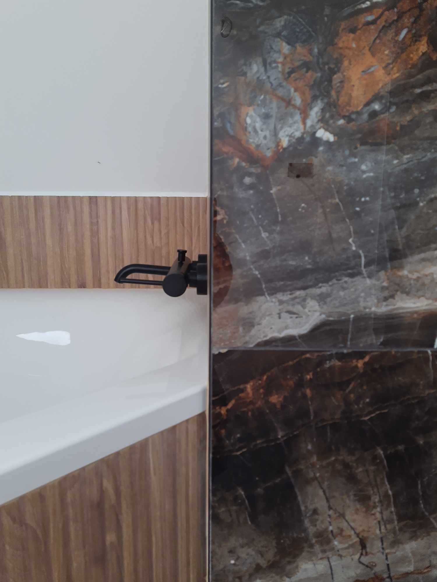 Fragment wykończonej łazienki: czarna bateria wannowa na tle drewnopodobnej okładziny i płytek imitujących marmur.