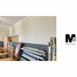 Wyremontowana sala lekcyjna z gładkimi, pomalowanymi ścianami w odcieniach beżu i szarości, widoczne biurka, szafki, tablica oraz zwinięte plansze i logo firmy Fach-Mat po prawej stronie.