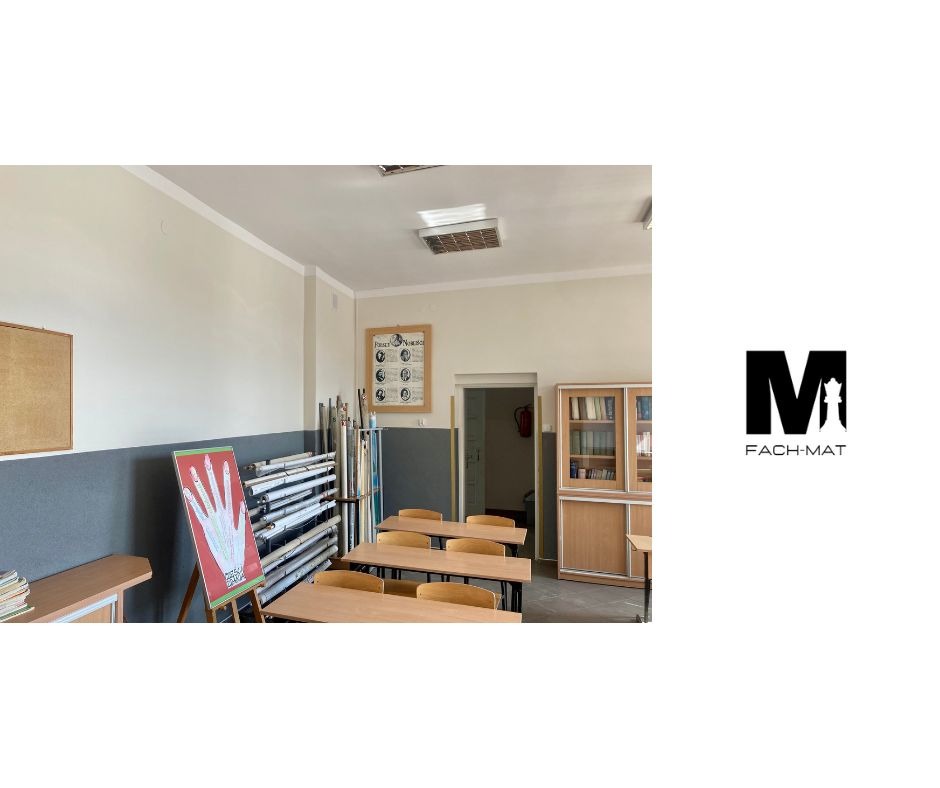 Sala lekcyjna z pomalowanymi ścianami na szaro i kremowo, z widoczną listwą przypodłogową, rzędem stołów i krzeseł, plakatem anatomicznym dłoni na sztaludze, tubami z materiałami budowlanymi...