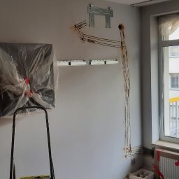 Energomax Sp. z o.o. - Montaż klimatyzacji w mieszkaniu: jednostka wewnętrzna w folii ochronnej na drabinie, widoczne przewody i uchwyty montażowe na ścianie, jednostka zewnętrzna montowana na balkonie przez montera.