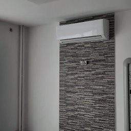 Energomax Sp. z o.o. - Biała klimatyzacja ścienna zamontowana na dekoracyjnej ścianie z szarej mozaiki w pomieszczeniu biurowym z tablicą interaktywną i projektorem.