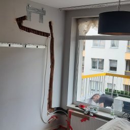 Energomax Sp. z o.o. - Montaż klimatyzacji w mieszkaniu: widoczne koryto w ścianie, uchwyt montażowy, przewody, pracownik zagląda przez okno na balkon.