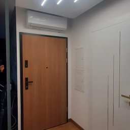 Energomax Sp. z o.o. - Nowoczesny przedpokój z klimatyzatorem nad brązowymi drzwiami wejściowymi, oświetlenie sufitowe LED, białe drzwi wewnętrzne z pionowymi frezami i drewniana podłoga