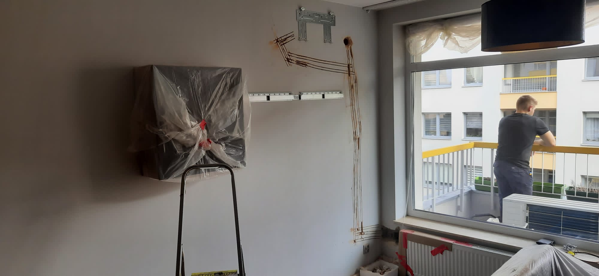 Montaż klimatyzacji w mieszkaniu: jednostka wewnętrzna w folii ochronnej na drabinie, widoczne przewody i uchwyty montażowe na ścianie, jednostka zewnętrzna montowana na balkonie przez montera.