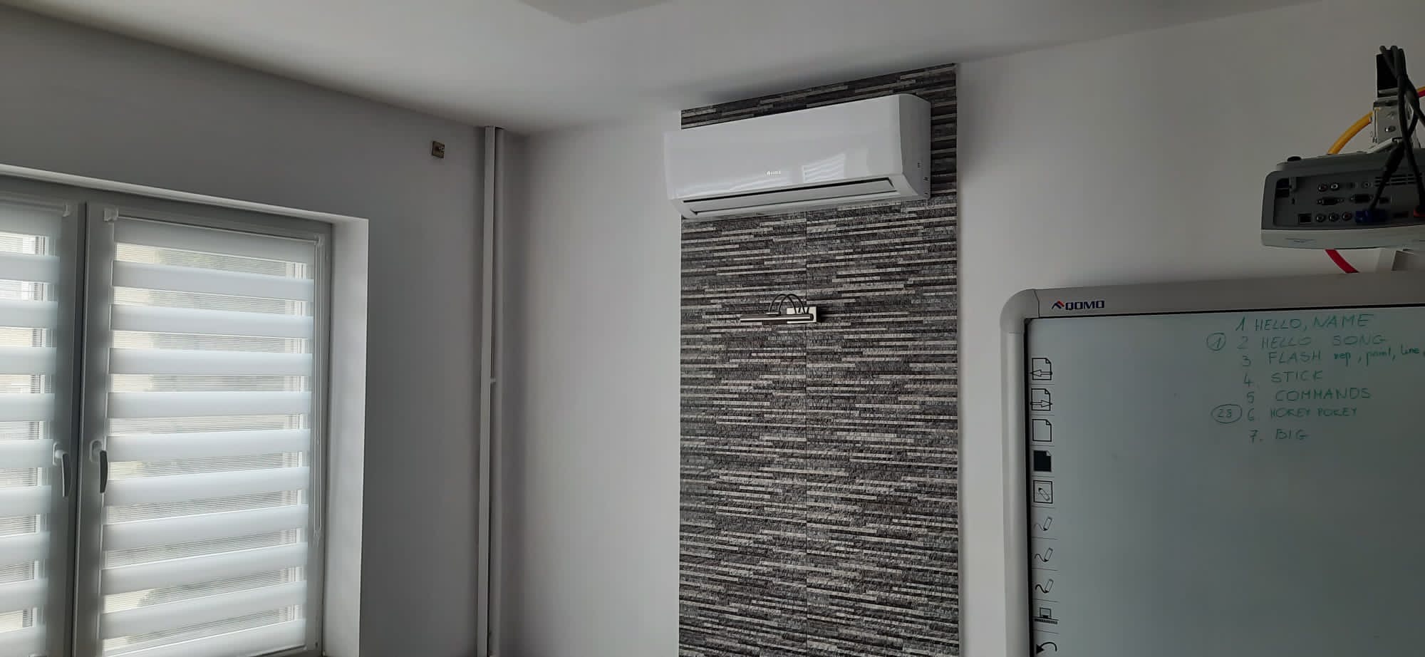 Biała klimatyzacja ścienna zamontowana na dekoracyjnej ścianie z szarej mozaiki w pomieszczeniu biurowym z tablicą interaktywną i projektorem.