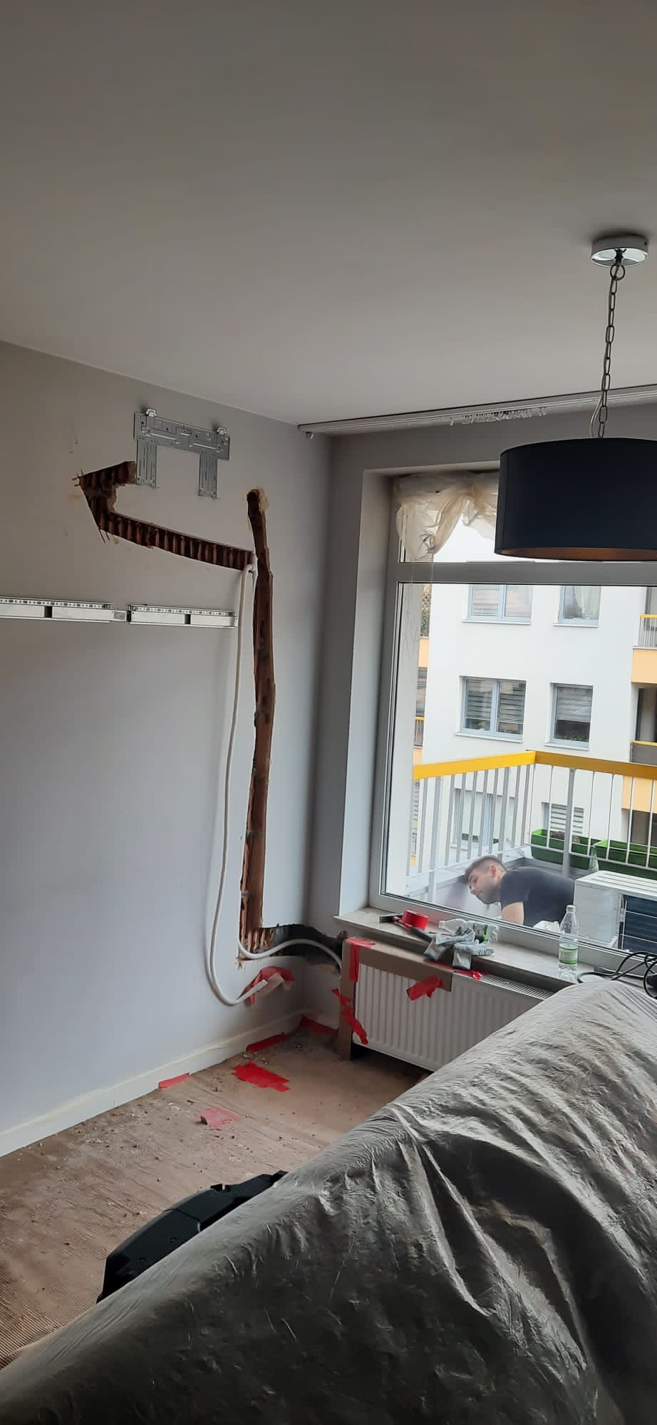 Montaż klimatyzacji w mieszkaniu: widoczne koryto w ścianie, uchwyt montażowy, przewody, pracownik zagląda przez okno na balkon.