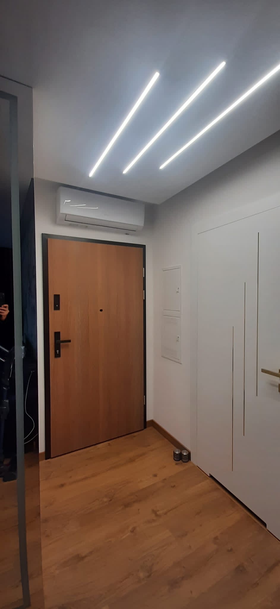 Nowoczesny przedpokój z klimatyzatorem nad brązowymi drzwiami wejściowymi, oświetlenie sufitowe LED, białe drzwi wewnętrzne z pionowymi frezami i drewniana podłoga