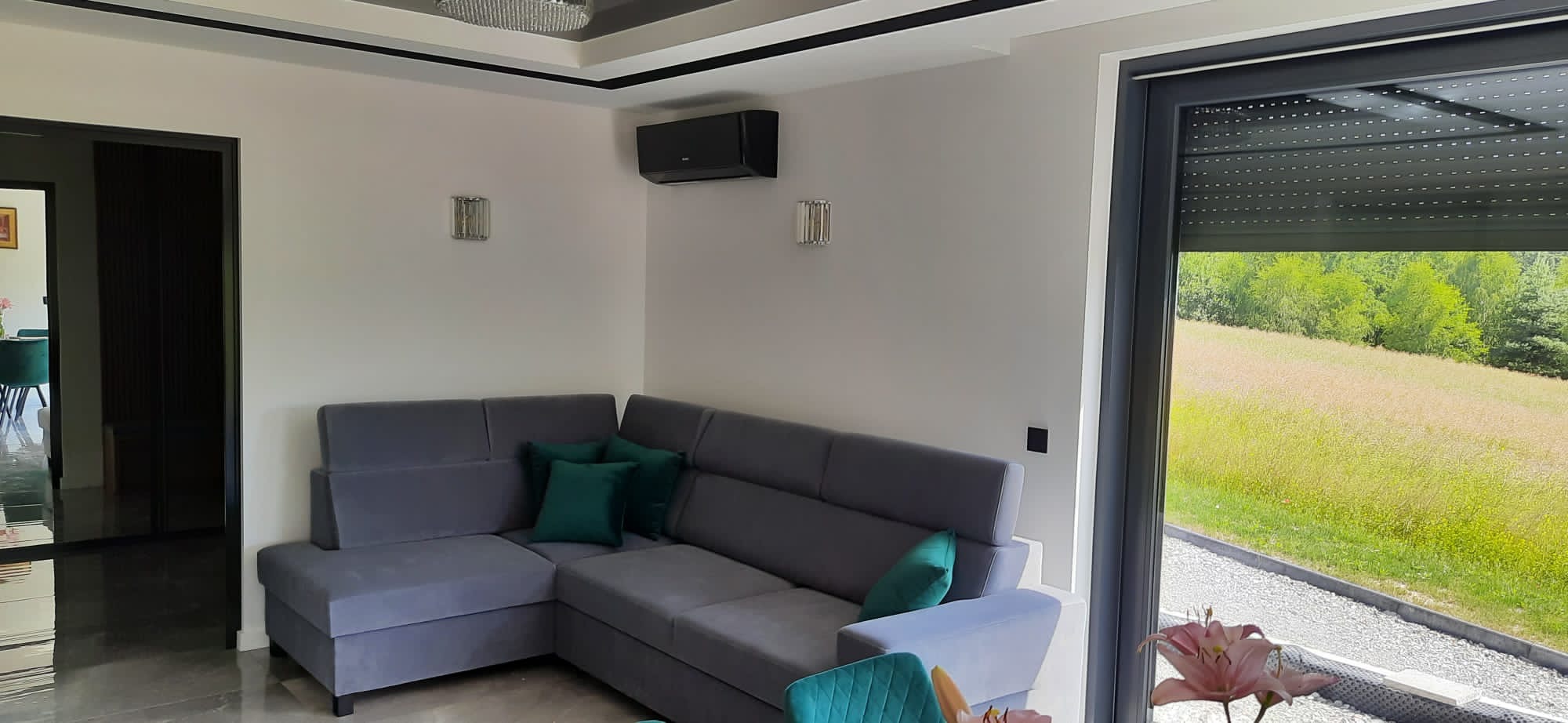 Nowoczesny salon z szarym narożnikiem, zielonymi poduszkami i czarną klimatyzacją ścienną, widok na łąkę przez okno z roletą.
