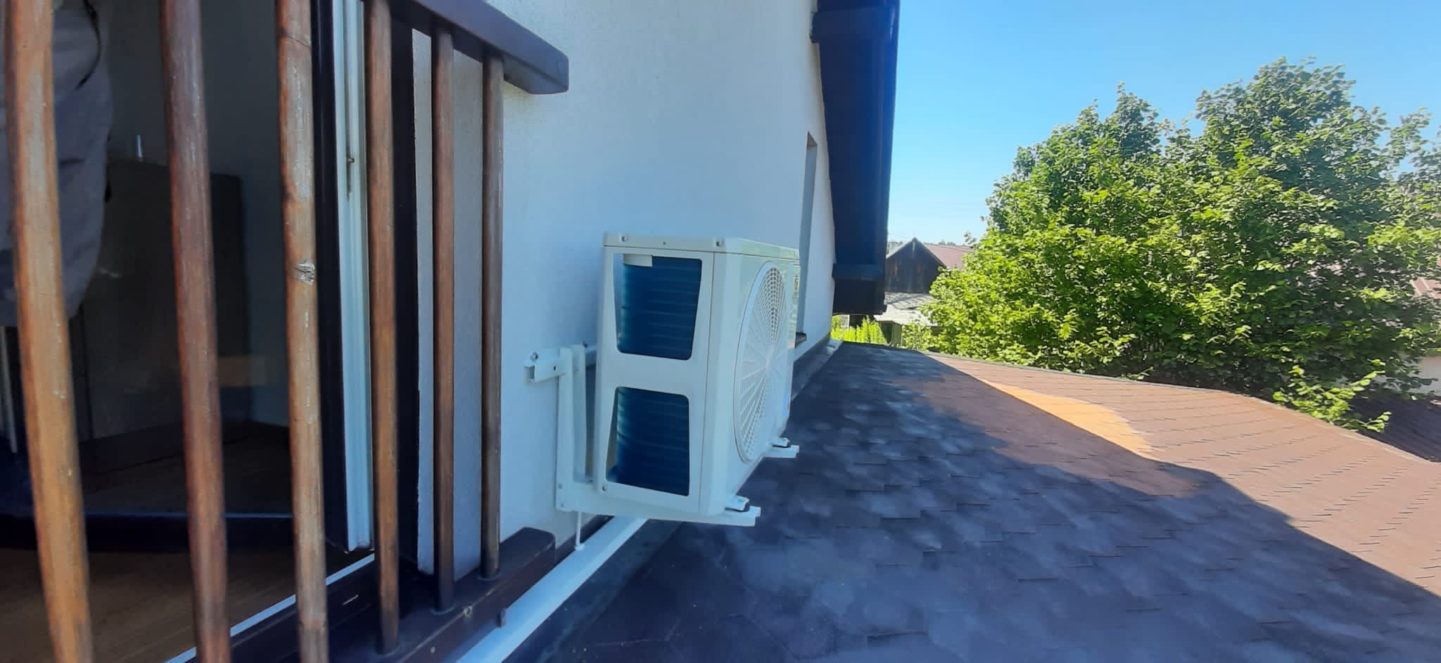 Biała jednostka zewnętrzna klimatyzacji zamontowana na ścianie domu, widoczna z perspektywy dachu, z drewnianą balustradą w kadrze i zielonym drzewem w tle.